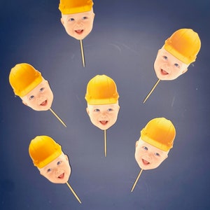 May include: A collection of baby face cutouts wearing yellow construction hats, attached to wooden skewers. The faces have blue eyes and are smiling. The background is a dark blue color.