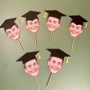 May include: Six black graduation cap cupcake toppers with different faces of people wearing the caps. The toppers are on a light green surface.