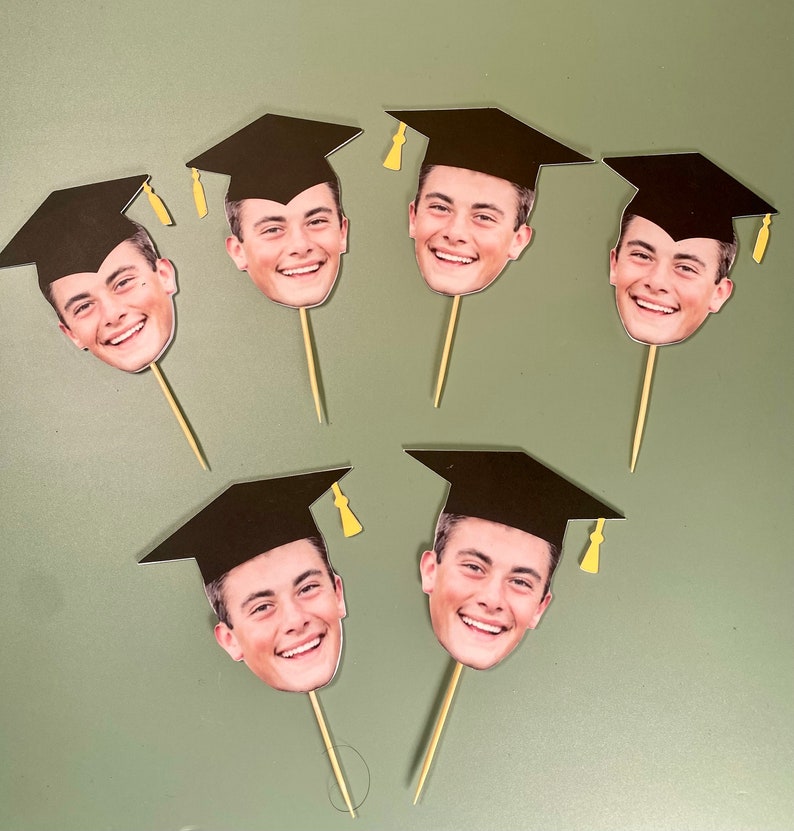 May include: Six black graduation cap cupcake toppers with different faces of people wearing the caps. The toppers are on a light green surface.