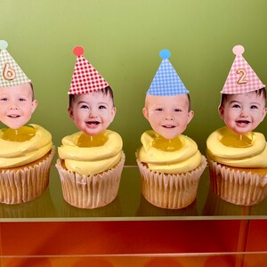 May include: Four cupcakes with yellow frosting and a white paper liner. Each cupcake has a different colored and patterned party hat with a child's face on top. The hats have the numbers 6 and 2 on them.