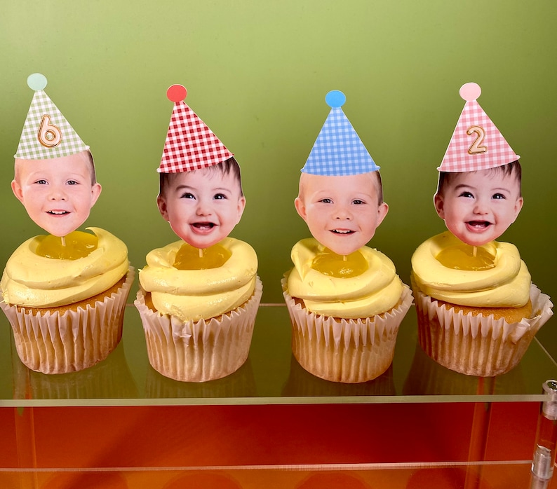 May include: Four cupcakes with yellow frosting and a white paper liner. Each cupcake has a different colored and patterned party hat with a child's face on top. The hats have the numbers 6 and 2 on them.
