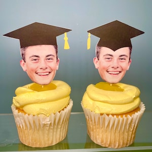 May include: Two yellow cupcakes with black graduation caps and faces on top. The cupcakes are on a glass surface.
