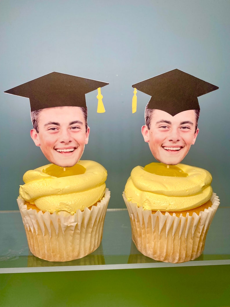 May include: Two yellow cupcakes with black graduation caps and faces on top. The cupcakes are on a glass surface.