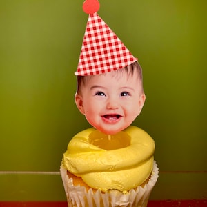 May include: A yellow cupcake with white frosting and a red and white checkered party hat with a red pom-pom on top. A baby's face is on the topper.