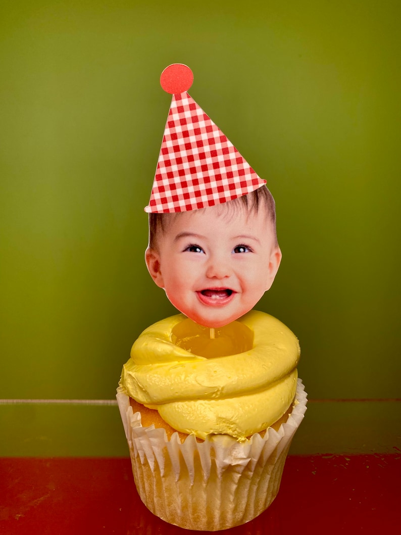 May include: A yellow cupcake with white frosting and a red and white checkered party hat with a red pom-pom on top. A baby's face is on the topper.