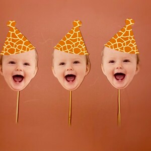 May include: Three orange and brown giraffe print party hats with a baby's face cut out in the front. The hats are on sticks.