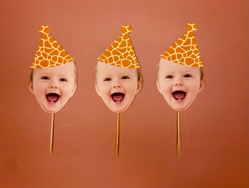 May include: Three orange and brown giraffe print party hats with a baby's face cut out in the front. The hats are on sticks.