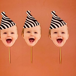 May include: Three black and white striped party hats with a child's face in each. The faces are smiling and have their mouths open. The hats are on sticks.