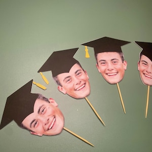 May include: Four graduation cap toppers with photos of people's faces. The caps are black with gold tassels. The toppers are on wooden skewers.