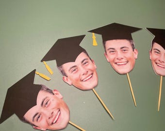 May include: Four graduation cap toppers with photos of people's faces. The caps are black with gold tassels. The toppers are on wooden skewers.