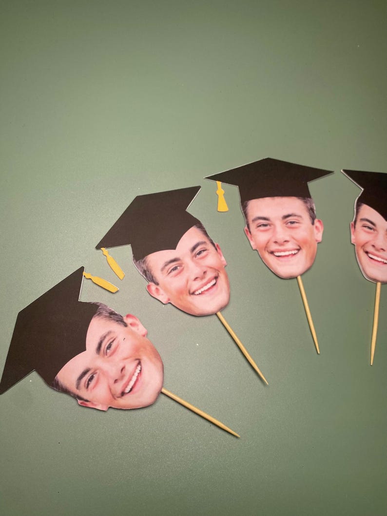 May include: Four graduation cap toppers with photos of people's faces. The caps are black with gold tassels. The toppers are on wooden skewers.