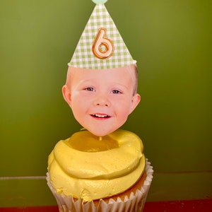 May include: A yellow cupcake with a green and white gingham party hat with a gold number six on it. A child's face is on a stick in the cupcake.