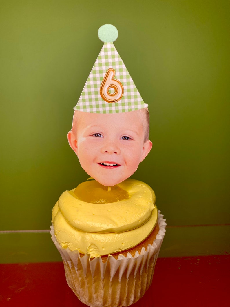 May include: A yellow cupcake with a green and white gingham party hat with a gold number six on it. A child's face is on a stick in the cupcake.