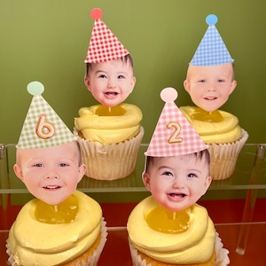 May include: Four yellow cupcakes with white frosting and a clear jelly center. Each cupcake has a different colored gingham party hat with a number on it and a photo of a baby's face on top.