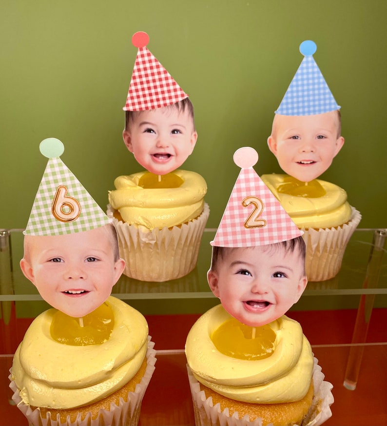 May include: Four yellow cupcakes with white frosting and a clear jelly center. Each cupcake has a different colored gingham party hat with a number on it and a photo of a baby's face on top.