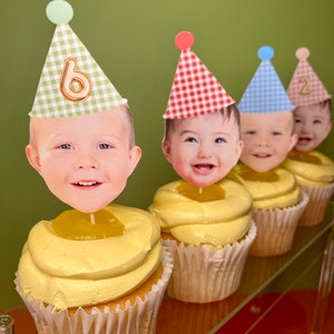 May include: Four cupcakes with yellow frosting and a lemon curd center. Each cupcake has a different colored party hat with a child's face and a number on it. The hats are green, red, blue, and pink. The numbers on the hats are 6, 1, 2, and 2.