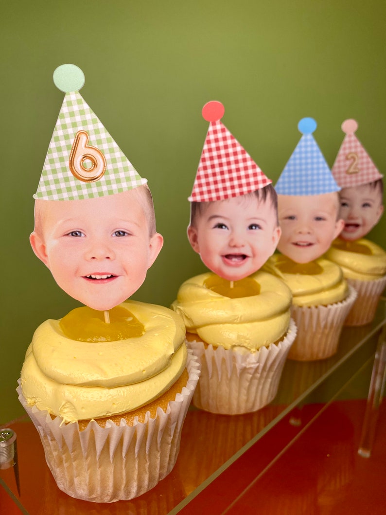 May include: Four cupcakes with yellow frosting and a lemon curd center. Each cupcake has a different colored party hat with a child's face and a number on it. The hats are green, red, blue, and pink. The numbers on the hats are 6, 1, 2, and 2.