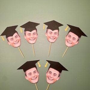 May include: Six black graduation caps with a face printed on each. The caps are on toothpicks and are arranged in a semi-circle.