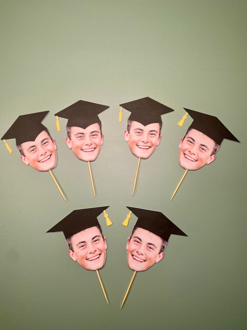 May include: Six black graduation caps with a face printed on each. The caps are on toothpicks and are arranged in a semi-circle.