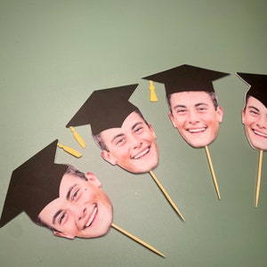 May include: Four graduation cap photo booth props with smiling faces. Each prop has a black graduation cap and a stick.