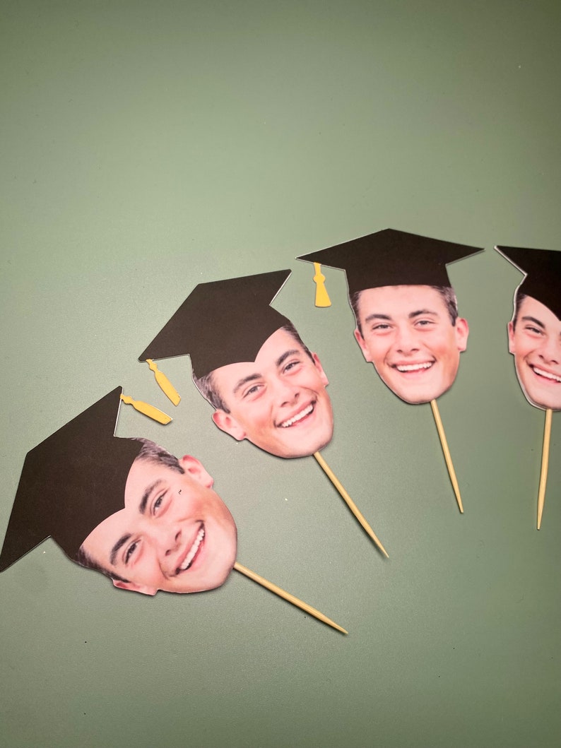 May include: Four graduation cap photo booth props with smiling faces. Each prop has a black graduation cap and a stick.