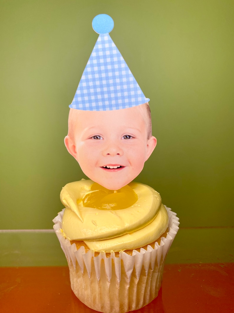 May include: A yellow cupcake with a blue and white checkered party hat and a smiling face cutout on top.
