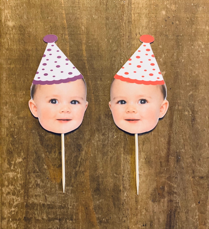 K&ouml;nnte beinhalten: Zwei Cupcake-Topper mit einem Babygesicht, das einen Partyhut tr&auml;gt. Die H&uuml;te sind wei&szlig; mit roten und lila Punkten. Die Topper sind auf Holzst&auml;bchen.