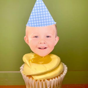 May include: A yellow cupcake with a white frosting swirl and a yellow jelly center. A blue and white checkered party hat with a blue pom-pom on top is on top of the cupcake. A child's face is on the front of the hat.