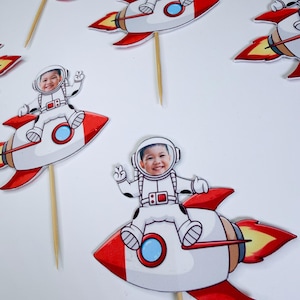 May include: Several cupcake toppers featuring a child in an astronaut suit riding a red, white, and brown rocket ship. The rocket has a blue circle and yellow flames. The toppers are attached to wooden sticks.