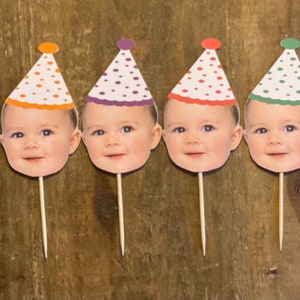K&ouml;nnte beinhalten: Sechs Cupcake-Topper mit einem Babygesicht und einer Partym&uuml;tze. Die M&uuml;tzen sind gelb, orange, lila, rot, gr&uuml;n und blau mit wei&szlig;en Punkten.