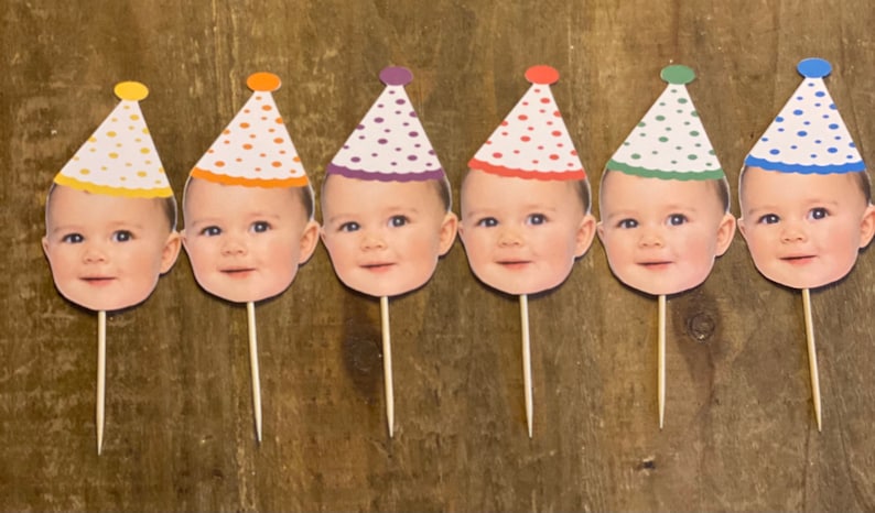 K&ouml;nnte beinhalten: Sechs Cupcake-Topper mit einem Babygesicht und einer Partym&uuml;tze. Die M&uuml;tzen sind gelb, orange, lila, rot, gr&uuml;n und blau mit wei&szlig;en Punkten.