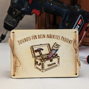 Può includere: Targa in legno chiaro con la scritta "Zuschuss für dein nächstes Projekt" e una grafica di cassetta degli attrezzi. La cassetta degli attrezzi contiene un martello e una sega. La targa è legata con spago.