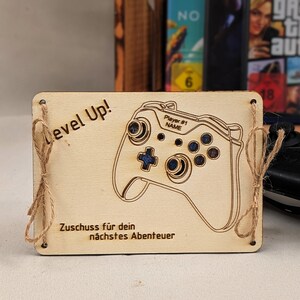 Könnte beinhalten: Helle Holzplakette mit einem gelaserten Design eines Videospiel-Controllers. Der Text "Level Up!" steht über dem Controller, und "Player #1 NAME" ist auf dem Controller geschrieben. Darunter steht "Zuschuss für dein nächstes Abenteuer".