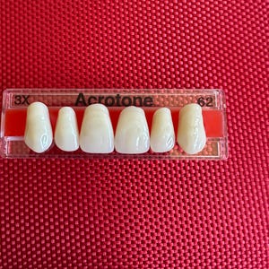 Puede incluir: Primer plano de un juego de seis dientes artificiales en una bandeja de plástico transparente. La bandeja está etiquetada con "3X AcriTone 62".