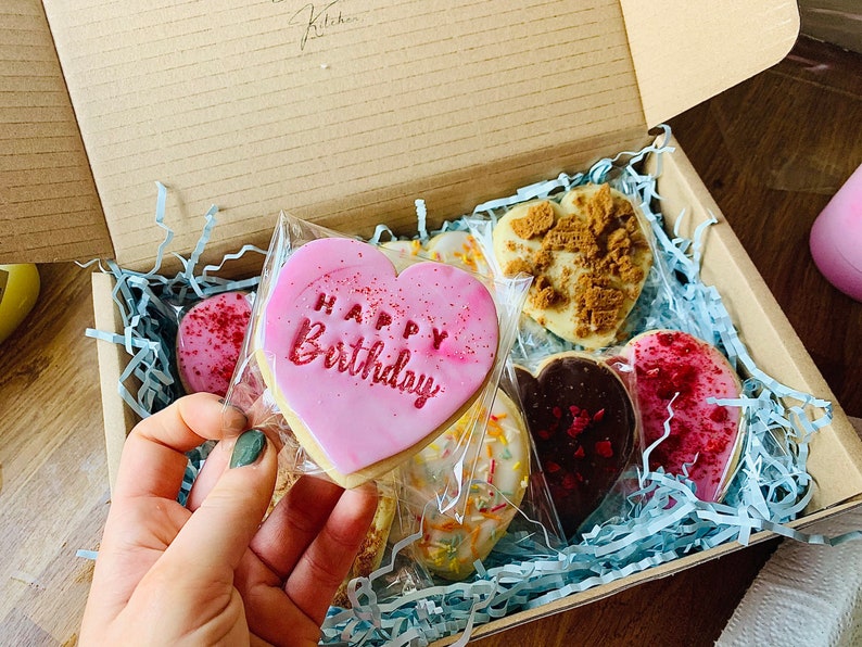 Puede incluir: Una caja de regalo llena de galletas en forma de coraz&oacute;n de color rosa, rojo y blanco. Una galleta tiene las palabras "Happy Birthday" escritas en glaseado rosa.