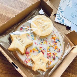 Pu&ograve; includere: Un biscotto bianco glassato con codette colorate e tre biscotti a forma di stella. I biscotti a forma di stella sono decorati con glassa dorata e le parole "Earl" e "50".