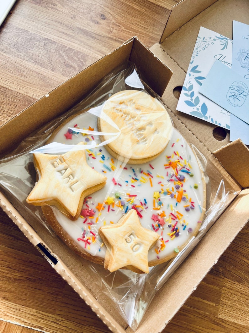 Pu&ograve; includere: Un biscotto bianco glassato con codette colorate e tre biscotti a forma di stella. I biscotti a forma di stella sono decorati con glassa dorata e le parole "Earl" e "50".