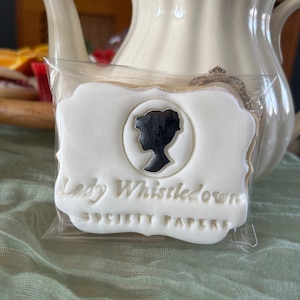 K&ouml;nnte beinhalten: Ein wei&szlig;er Keks mit einer schwarzen Silhouette eines Frauenkopfes und dem Text "Lady Whistledown Society Papers" in goldenen Buchstaben.
