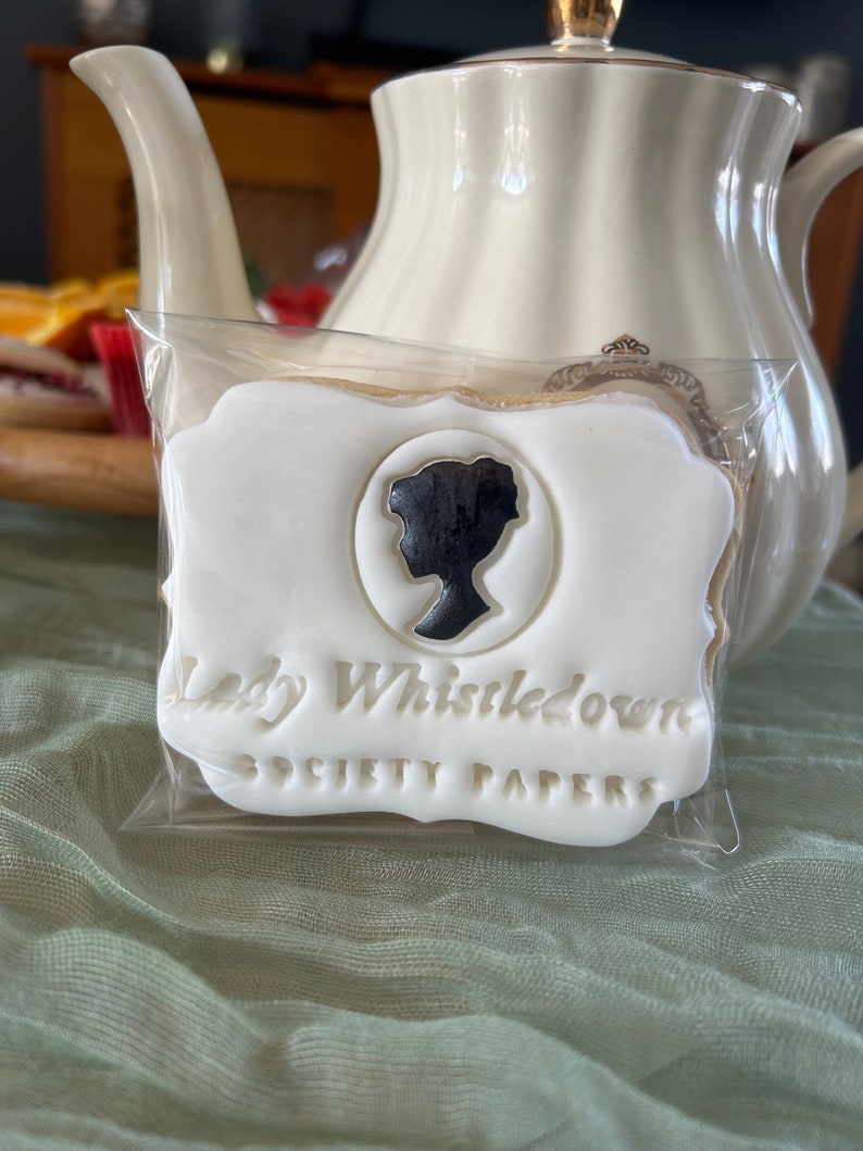 K&ouml;nnte beinhalten: Ein wei&szlig;er Keks mit einer schwarzen Silhouette eines Frauenkopfes und dem Text "Lady Whistledown Society Papers" in goldenen Buchstaben.