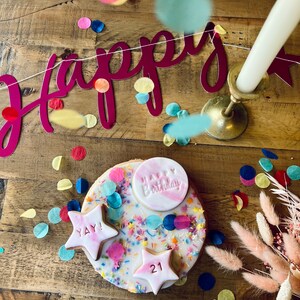 Pu&ograve; includere: Una torta bianca decorata con codette colorate, coriandoli e glassa rosa. La torta ha una stella rosa con il numero 21, una stella rosa con le parole "YAY!" e un cerchio rosa con le parole "Happy Birthday". La torta &egrave; circondata da coriandoli colorati e uno striscione rosa e bianco che dice "Happy".