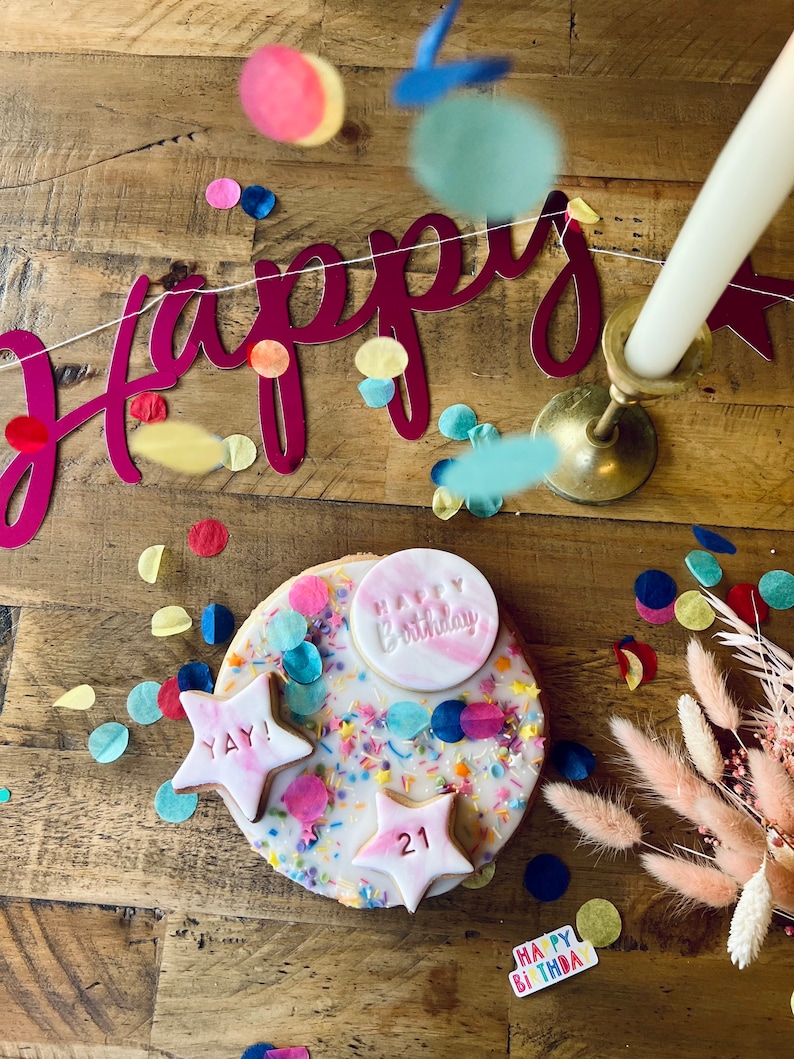 Pu&ograve; includere: Una torta bianca decorata con codette colorate, coriandoli e glassa rosa. La torta ha una stella rosa con il numero 21, una stella rosa con le parole "YAY!" e un cerchio rosa con le parole "Happy Birthday". La torta &egrave; circondata da coriandoli colorati e uno striscione rosa e bianco che dice "Happy".