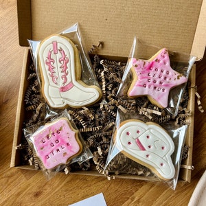 May include: A set of four pink and white decorated sugar cookies in a brown cardboard box. The cookies are shaped like a cowboy boot, a star, a square, and a cowboy hat. The star cookie has the text "Long Live Cowgirls" on it. The square cookie has the text "Let's Go Girls" on it.