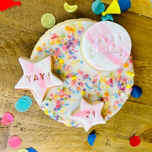 Pu&ograve; includere: Un biscotto rotondo di zucchero decorato con glassa bianca, codette colorate e stelle di pasta di zucchero rosa. Una stella dice "YAY!" e l'altra dice "21". Un cerchio di pasta di zucchero rosa pi&ugrave; grande dice "Buon compleanno".