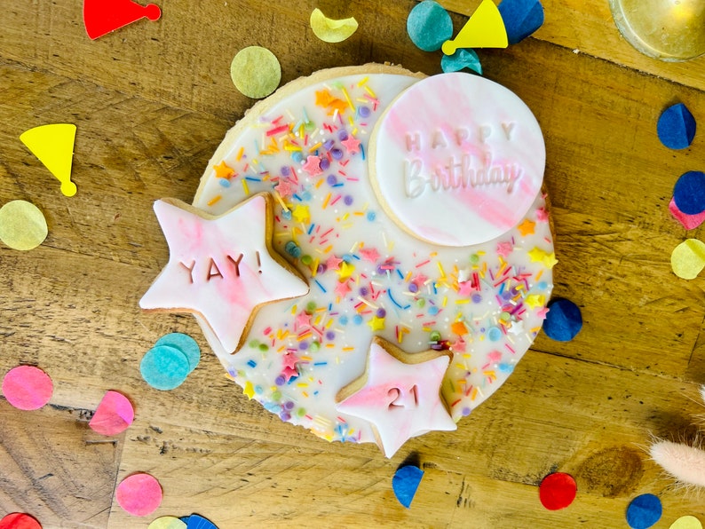 Pu&ograve; includere: Un biscotto rotondo di zucchero decorato con glassa bianca, codette colorate e stelle di pasta di zucchero rosa. Una stella dice "YAY!" e l'altra dice "21". Un cerchio di pasta di zucchero rosa pi&ugrave; grande dice "Buon compleanno".