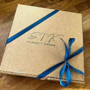 Pu&ograve; includere: Una scatola di cartone marrone con un nastro blu legato intorno. La scatola ha il testo "ETH Georgian Inspired" stampato su di essa.