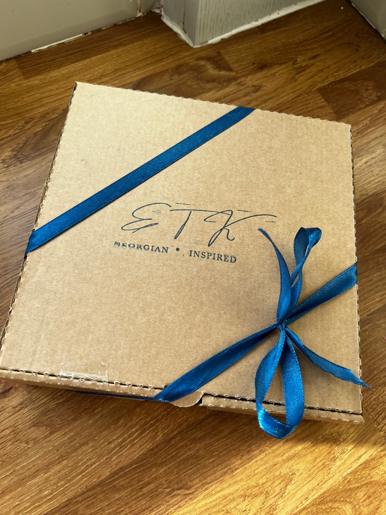 Pu&ograve; includere: Una scatola di cartone marrone con un nastro blu legato intorno. La scatola ha il testo "ETH Georgian Inspired" stampato su di essa.