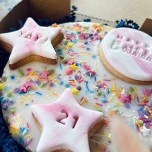Pu&ograve; includere: Una torta rotonda glassata di bianco decorata con biscotti a forma di stella rosa e bianca, spolverata con confettini colorati e il testo "Buon compleanno".