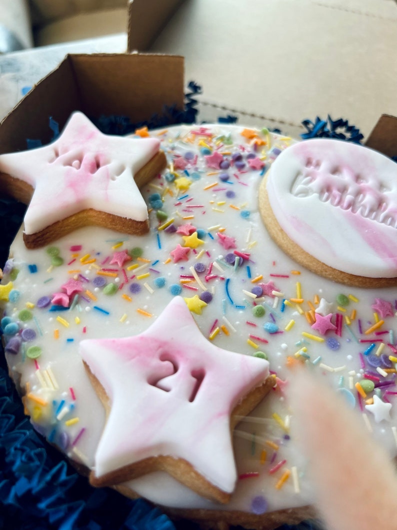 Pu&ograve; includere: Una torta rotonda glassata di bianco decorata con biscotti a forma di stella rosa e bianca, spolverata con confettini colorati e il testo "Buon compleanno".