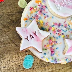 Pu&ograve; includere: Un biscotto rotondo bianco glassato e decorato con codette colorate e due biscotti di pasta di zucchero rosa e bianco. Un biscotto dice "Buon compleanno" e l'altro dice "Evviva!"