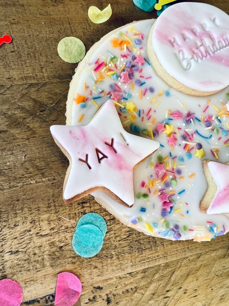 Pu&ograve; includere: Un biscotto rotondo bianco glassato e decorato con codette colorate e due biscotti di pasta di zucchero rosa e bianco. Un biscotto dice "Buon compleanno" e l'altro dice "Evviva!"